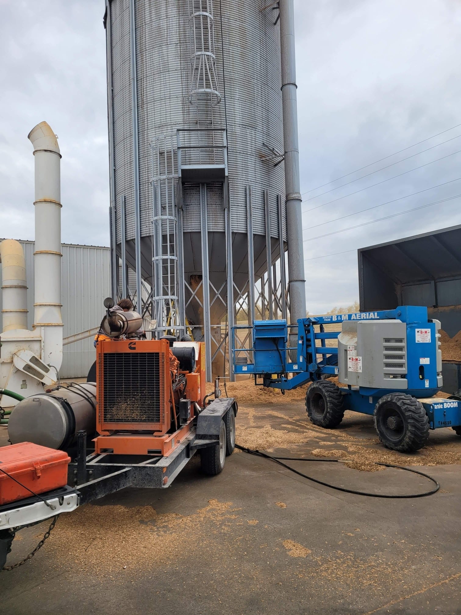 Large grain bin with industrial equipment, including a generator and aerial lift, in an outdoor setting.