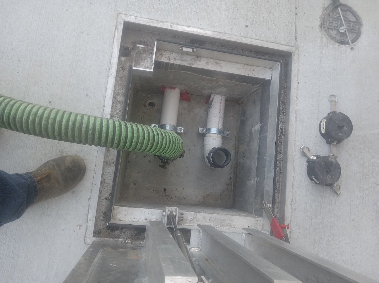 Green hose connected to a drain pipe inside a concrete pit with a boot in the foreground.