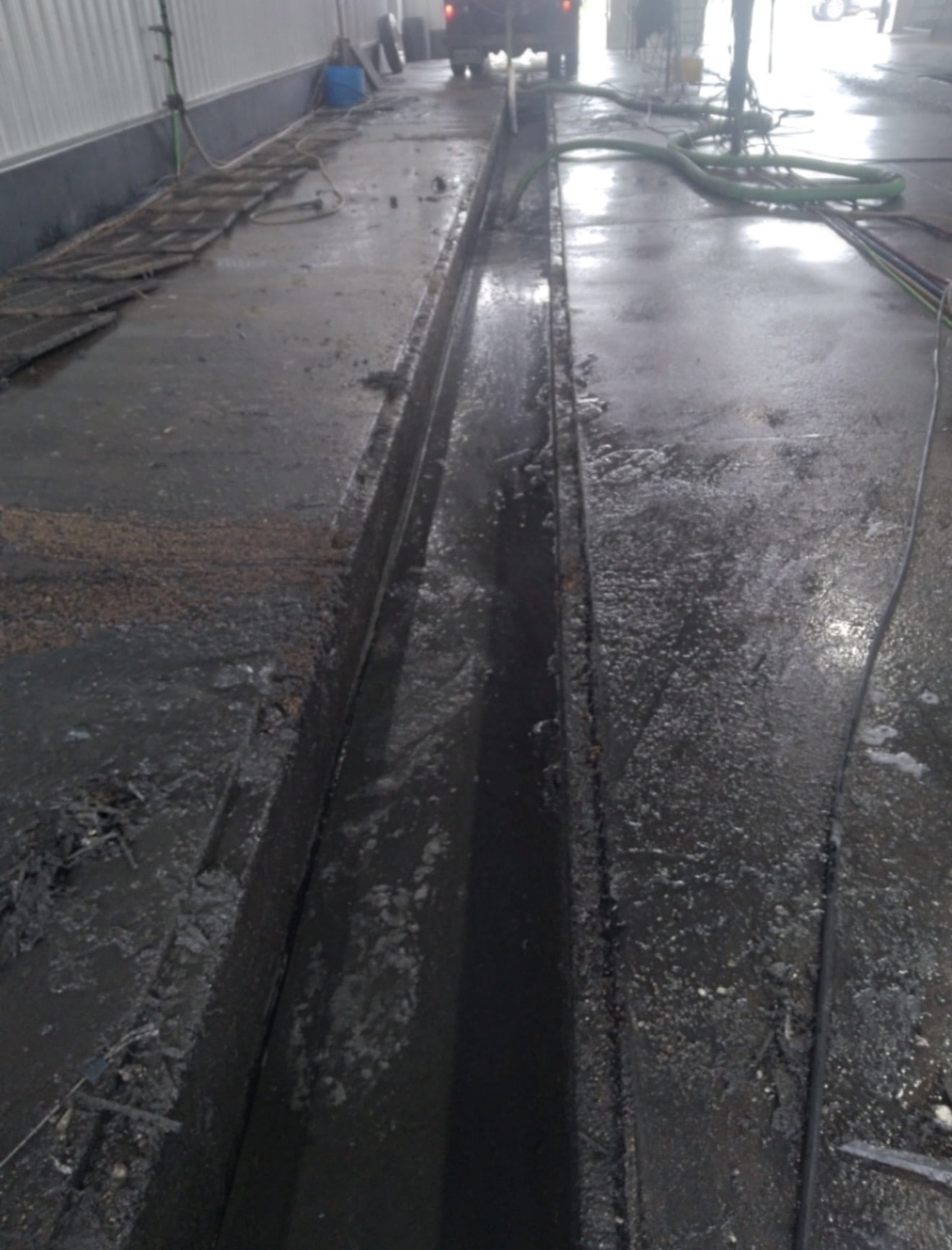 Grimy car wash pit with debris, puddles, and green hoses, wet concrete floor.